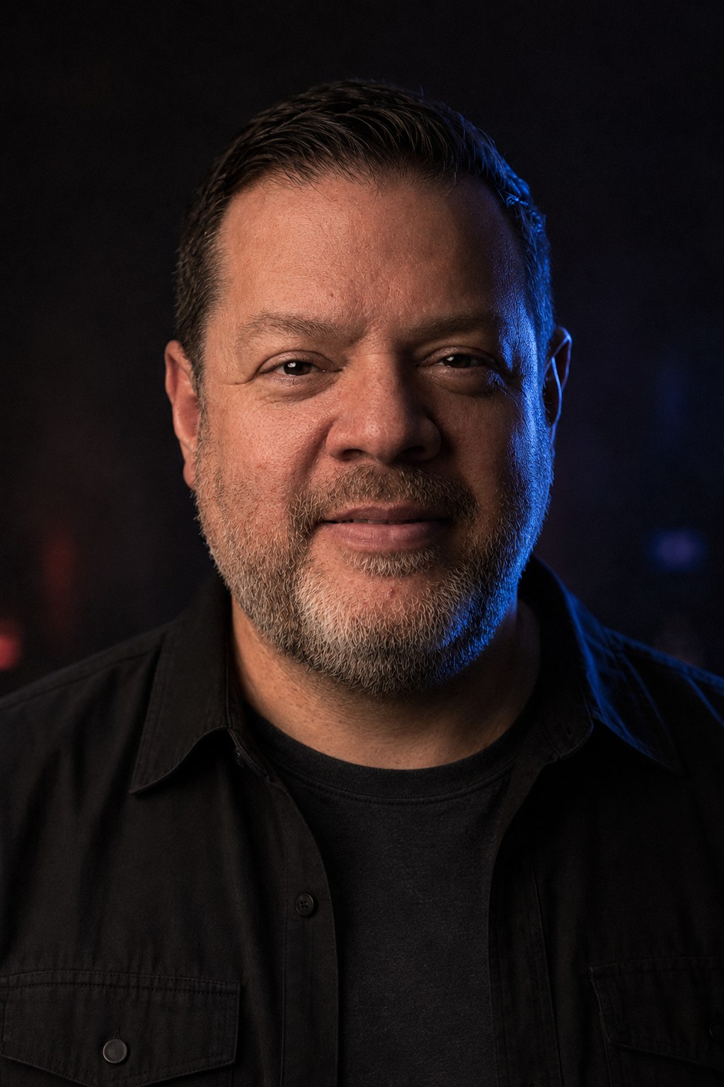 Professional headshot of Glenn Frank, live event saxophonist, wearing a black collared shirt against a dark background with blue rim lighting